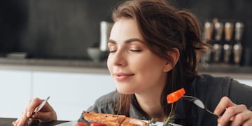 Image of pretty young woman sitting in kitchen while eating and smells fish and tomatoes\u002E Eyes closed\u002E