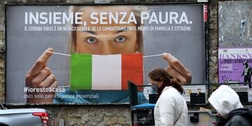 People walk past a huge billboard that shows a woman with as protective mask in the colours of an Italian flag and which reads, \